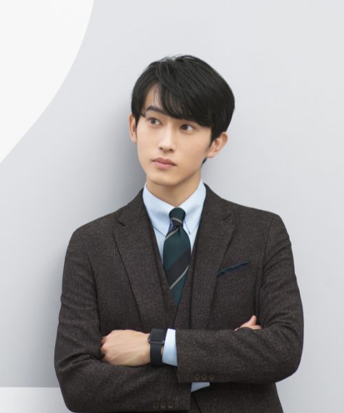杉野さんが腕組をして、淡いグレーの壁面に寄りかかっている。
ブラウン系のスリーピーススーツスタイルで、淡いブルー系のシャツ、グリーンとネイビー系の大きなストライプ柄のネクタイを合わせている。チーフもグリーン系。黒い腕時計を着けている。
上半身のカット。右斜め前方を見据えている。